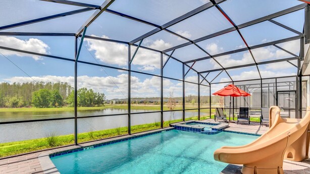 A covered outdoor swimming pool with a slide, surrounded by a metal frame and glass panels, overlooking a lake and greenery.