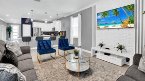 A modern living room with a glass coffee table, blue velvet chairs, a white entertainment unit, and a beach-themed TV wall.