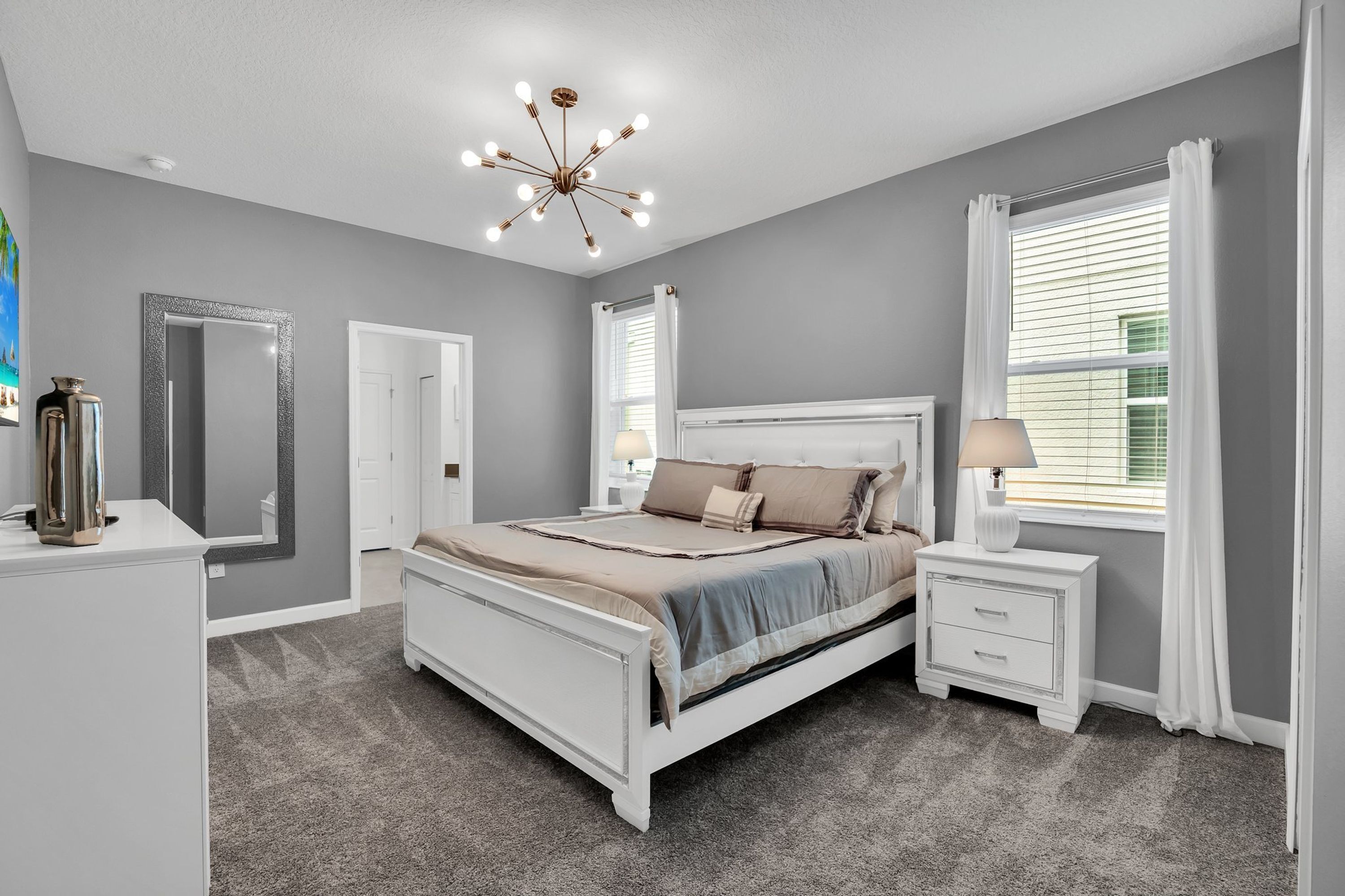 A bedroom with a bed, bedside tables, a mirror, a chandelier, and a window with curtains.