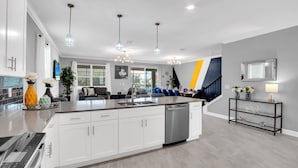 A modern kitchen with white cabinets, a central island, stainless steel appliances, pendant lighting, and a staircase leading to a dining area.