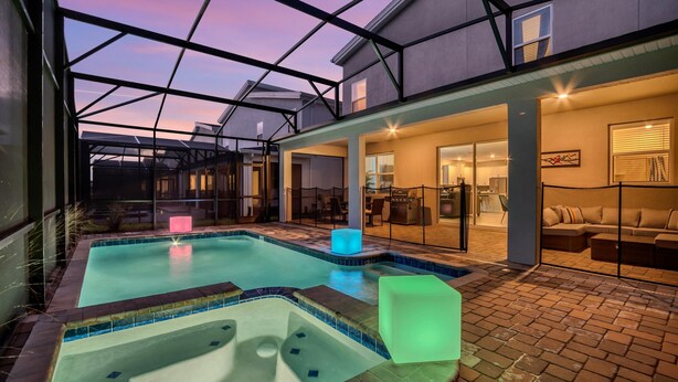A swimming pool with a hot tub, a glass-enclosed patio, and a well-lit interior space with a sofa and dining area.