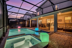 A swimming pool with a hot tub, a glass-enclosed patio, and a well-lit interior space with a sofa and dining area. - Amazing Family Home 8min to Disneyworld Pool Spa Sleeps 24 Stl101 (Kissimmee)