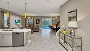 A modern kitchen with a central island, a dining area with a glass table, and a living room with a sofa and TV. - Magical Family Home 8min-disneyworld Pool Spa Sleeps 22 Stl103 (Kissimmee)