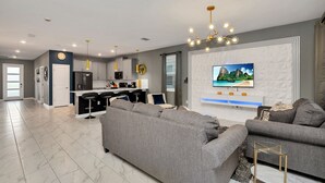 A modern living room with a grey sofa, a TV mounted on the wall, and a kitchen in the background. - Disney Decorated Home 8min to Disneyworld Pool Spa Sleeps 22 Stl104 (Kissimmee)