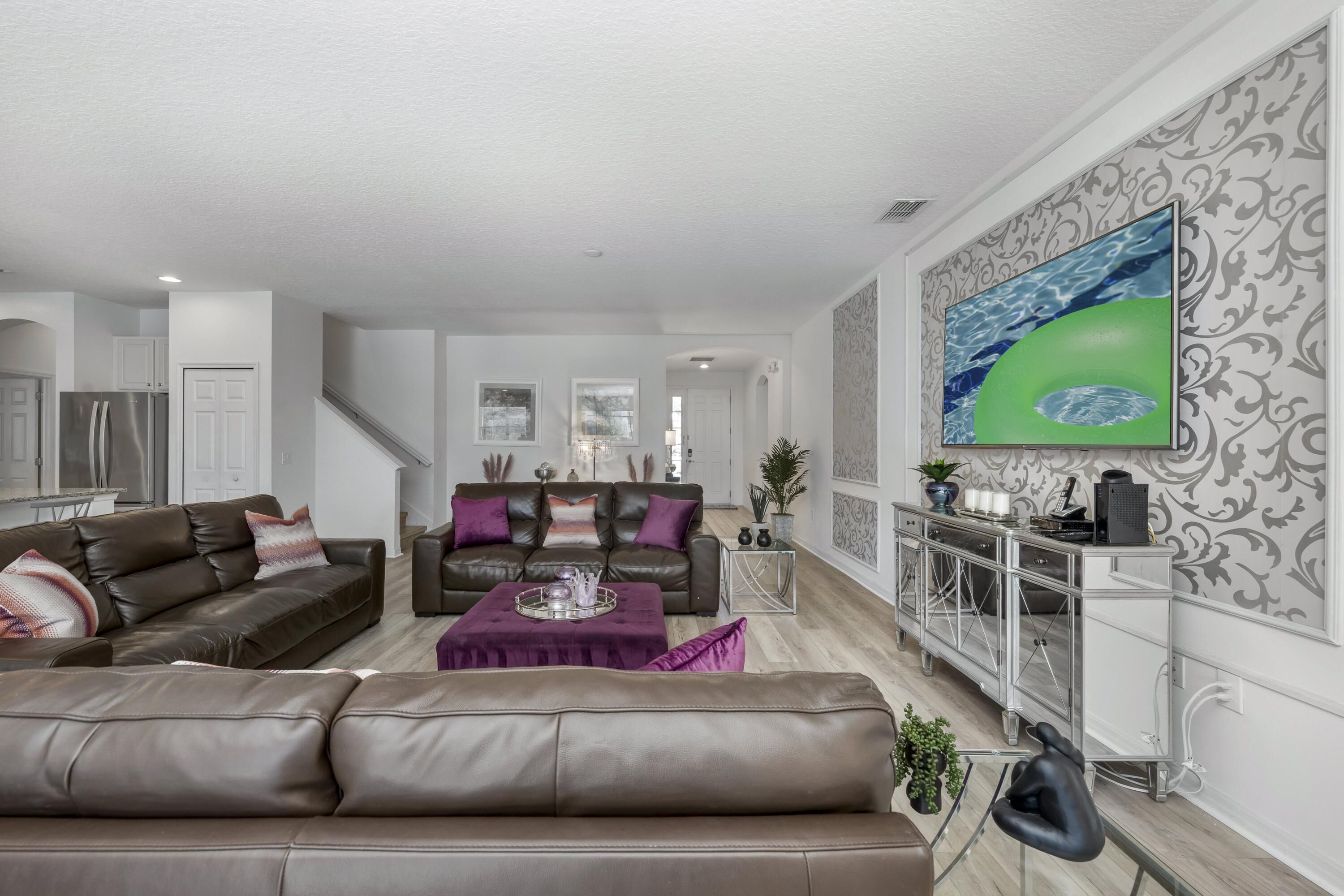 A spacious living room with a brown leather sofa, a matching armchair, and a coffee table. A dining area with a glass cabinet and a painting is visible in the background.