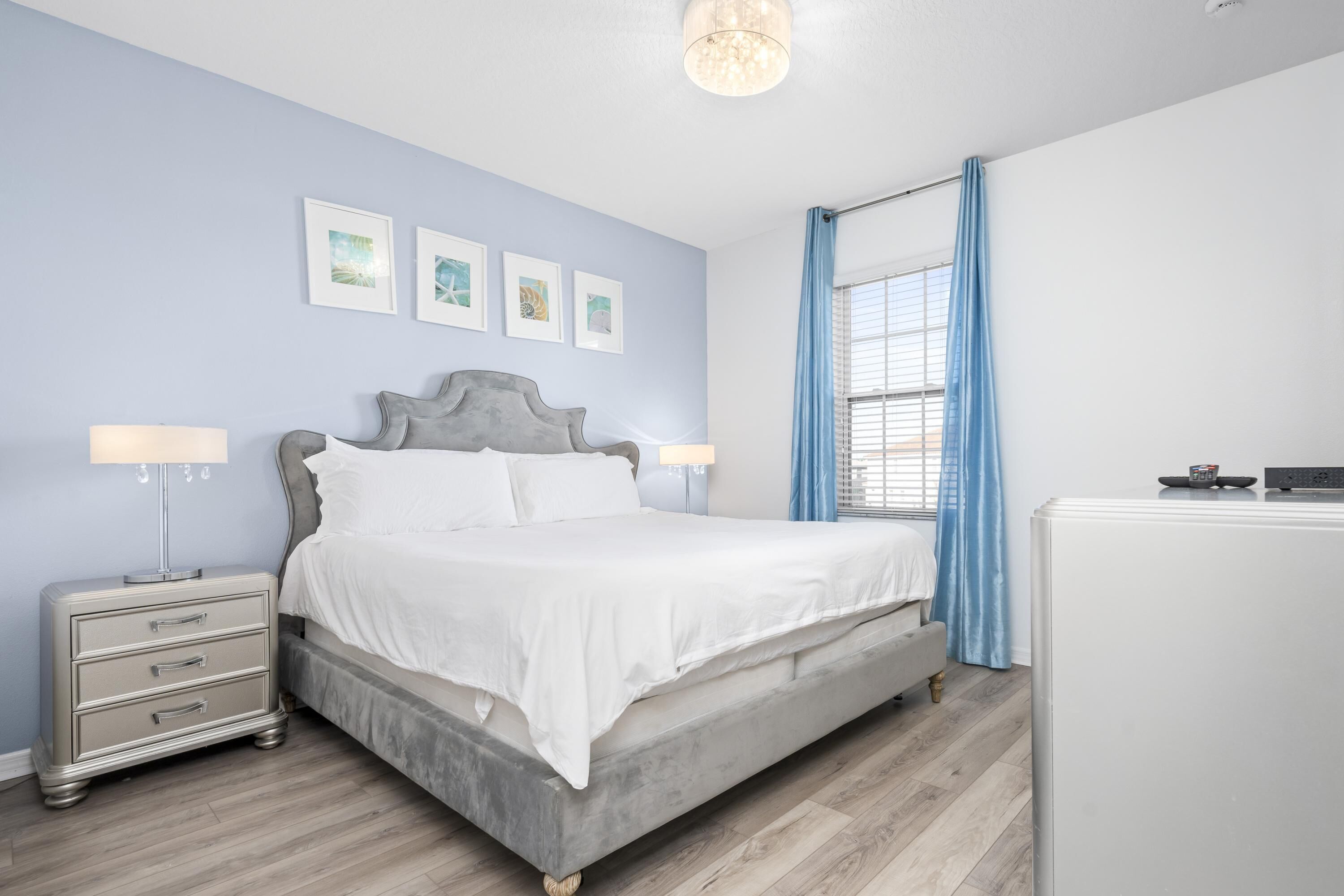 A bedroom with a bed, bedside tables, a nightstand, a dresser, a window with curtains, and wall art.