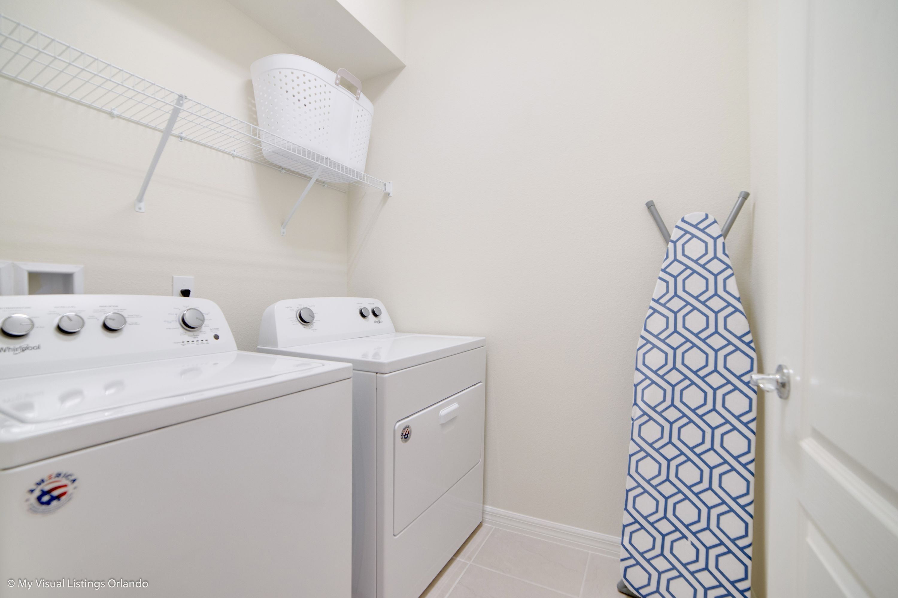 A laundry area with two front-loading washers and dryers, a white basket on a wire shelf, and a door with a patterned curtain.