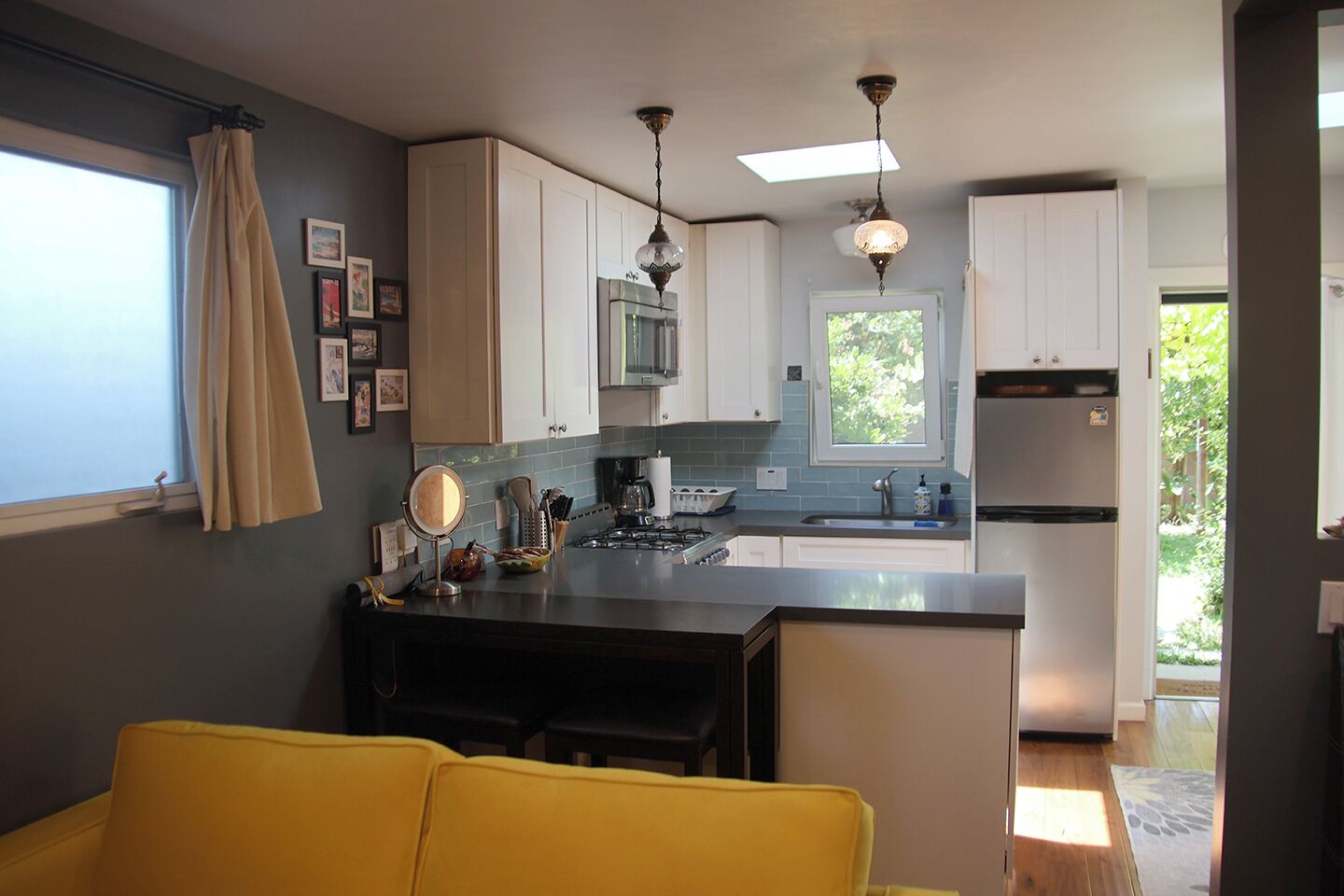 Une cuisine compacte avec des meubles blancs, une cuisinière à gaz, un évier et un réfrigérateur. On y trouve également un coin repas avec une chaise jaune et une petite table.