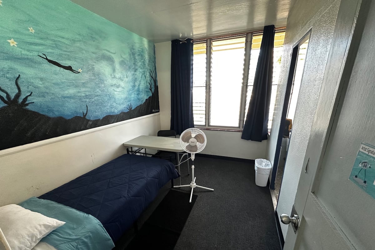 A compact room with a bed, a small table, a standing fan, and a window with curtains.