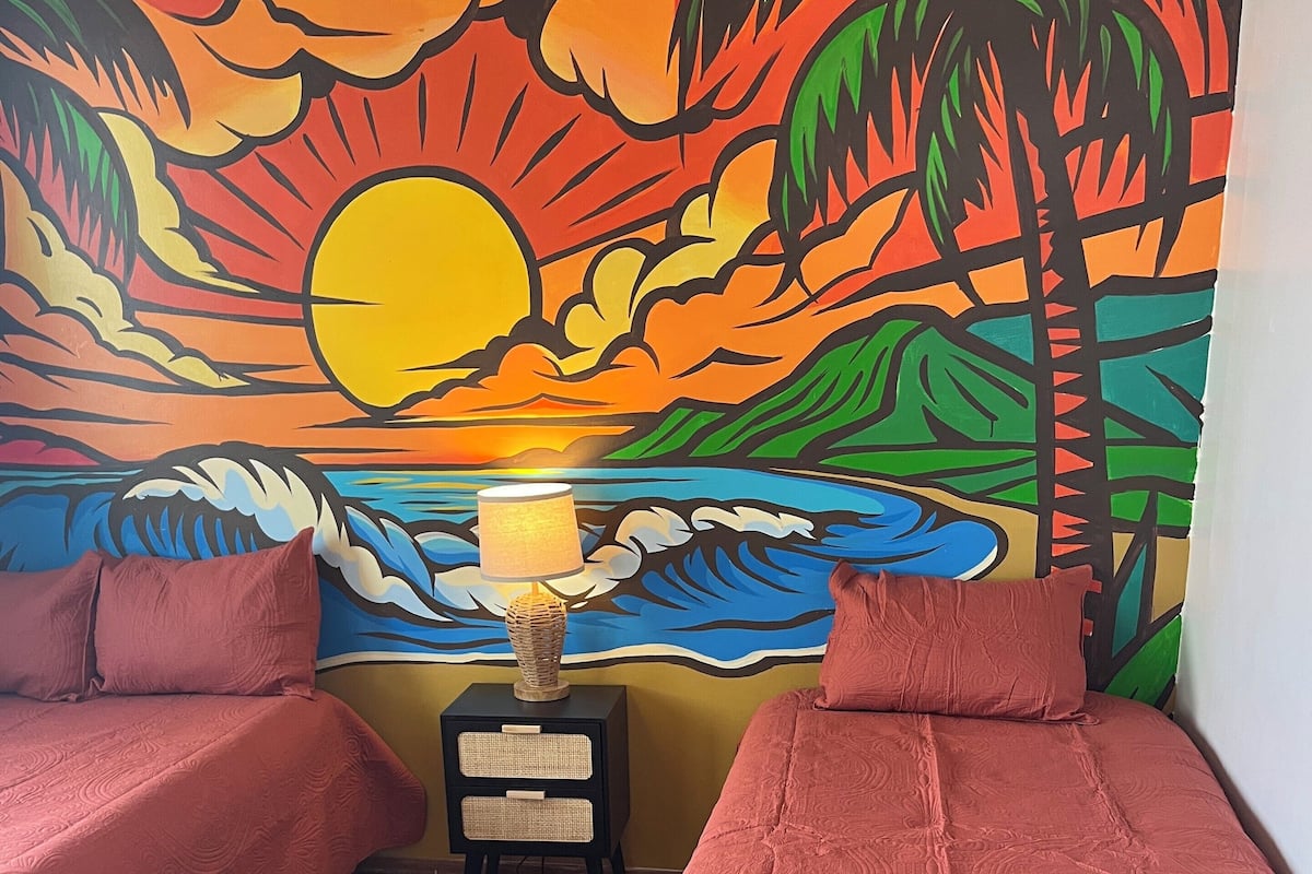 A room with two beds, a nightstand with a lamp, and a colorful mural on the wall.