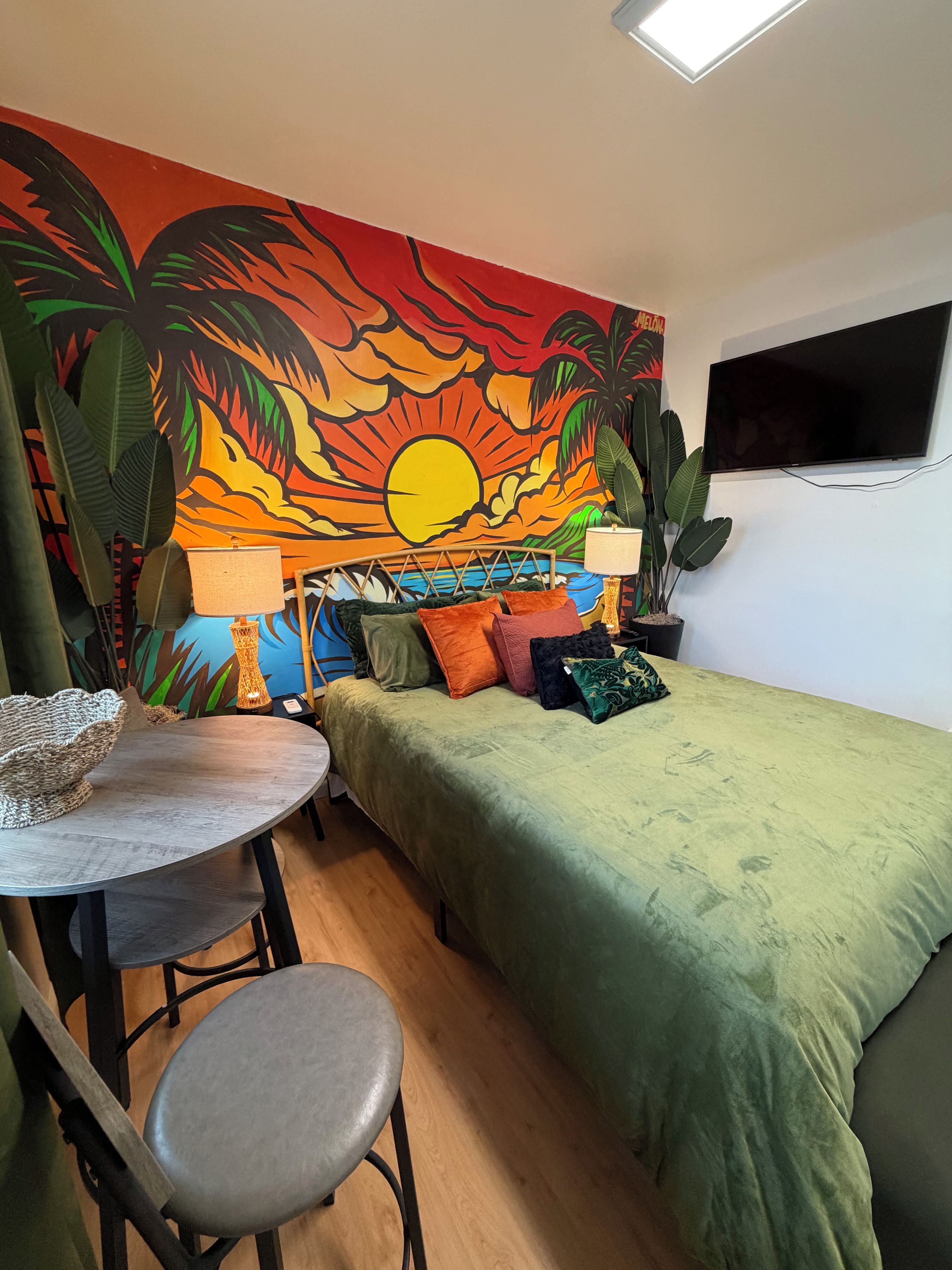 Une pièce comprenant un lit, une table, des chaises, un téléviseur et une fresque représentant un coucher de soleil.