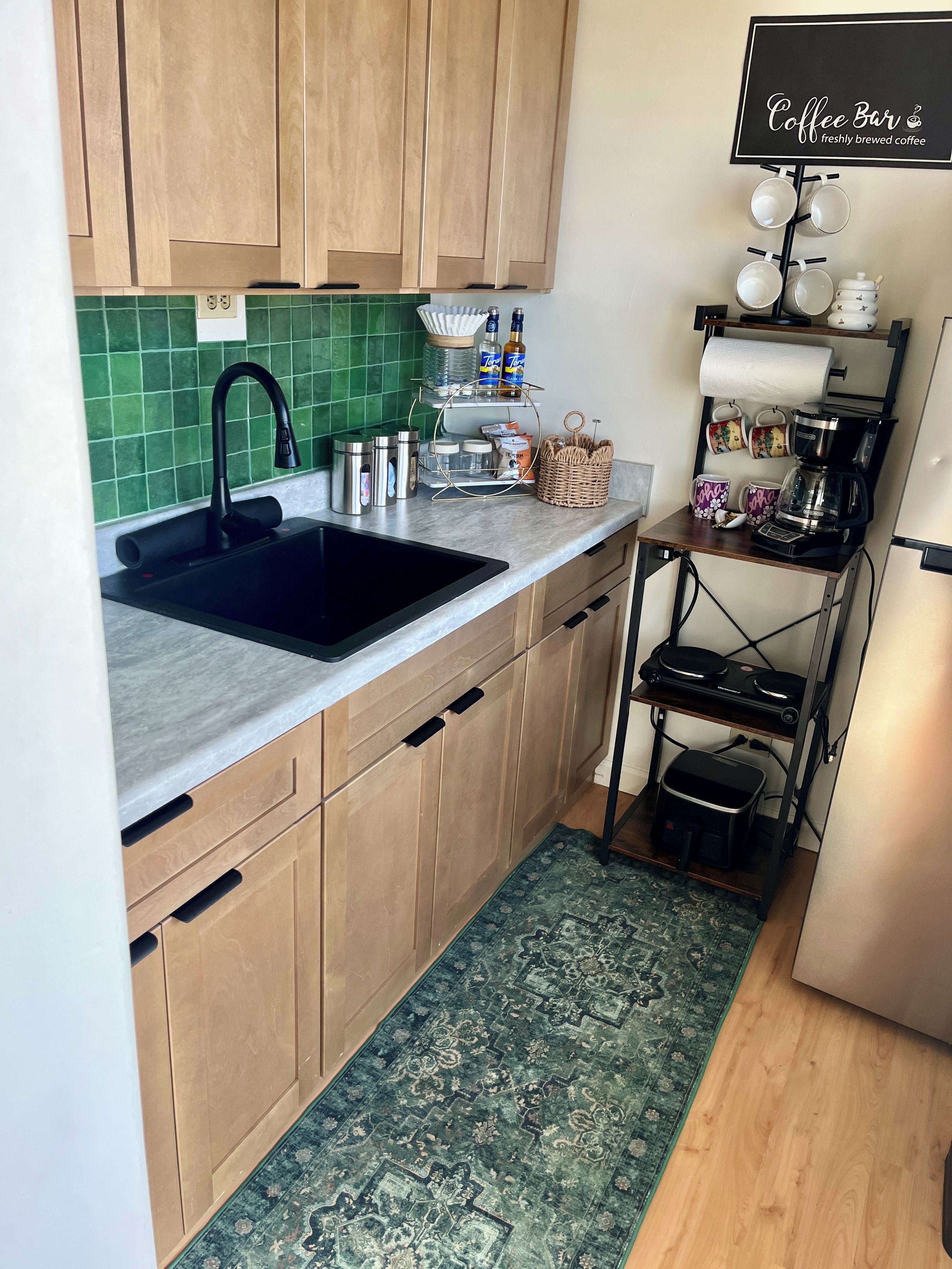 A kitchen with a black sink, wooden cabinets, a green tiled backsplash, a coffee station with mugs and a coffee maker, and a patterned rug.
