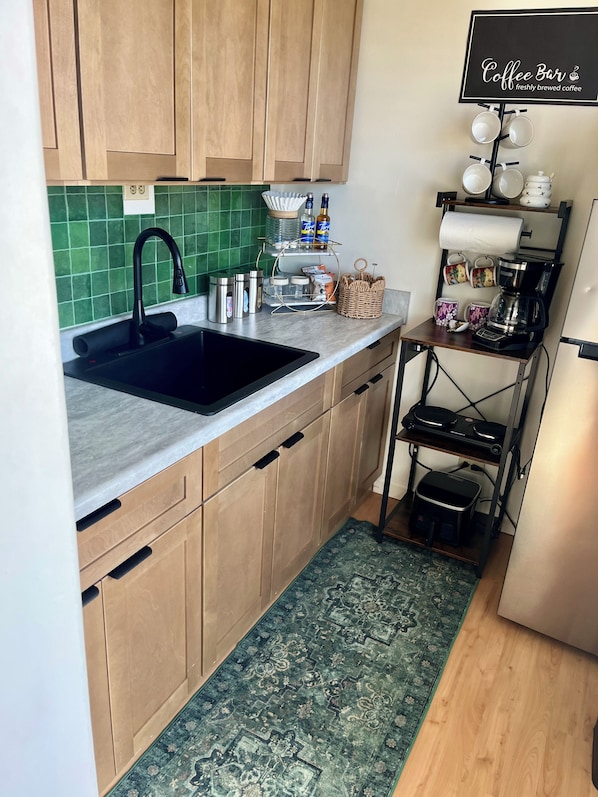 A kitchen with a black sink, wooden cabinets, a green tiled backsplash, a coffee station with mugs and a coffee maker, and a patterned rug. - Central YMCA Residences (Honolulu)