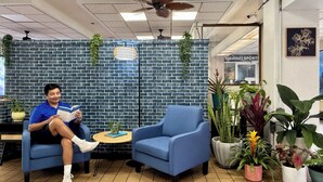 A man sitting on a blue chair reading a book in a room with a brick wall, a ceiling fan, and various potted plants. - Central YMCA Residences (Honolulu)