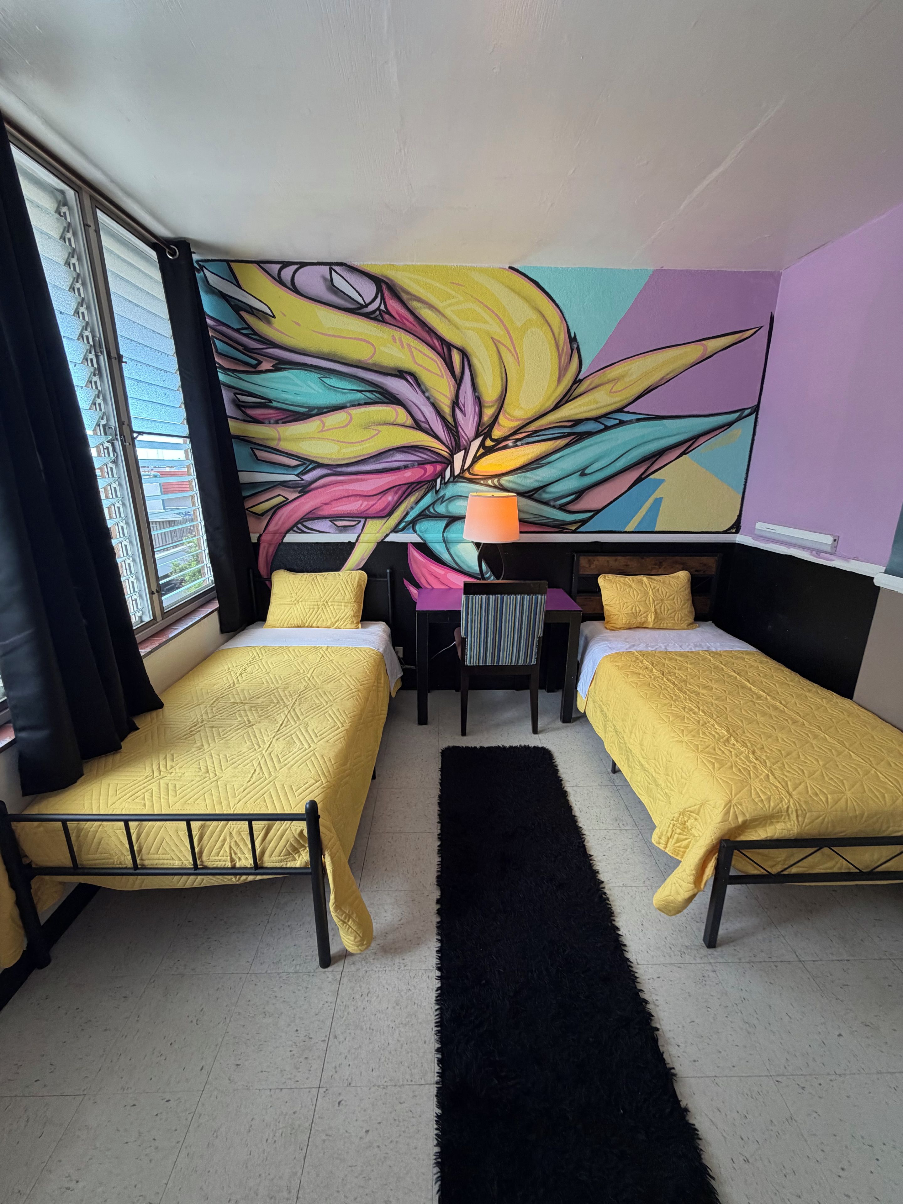 Una habitación con dos camas, un escritorio y un mural grande y colorido en la pared.