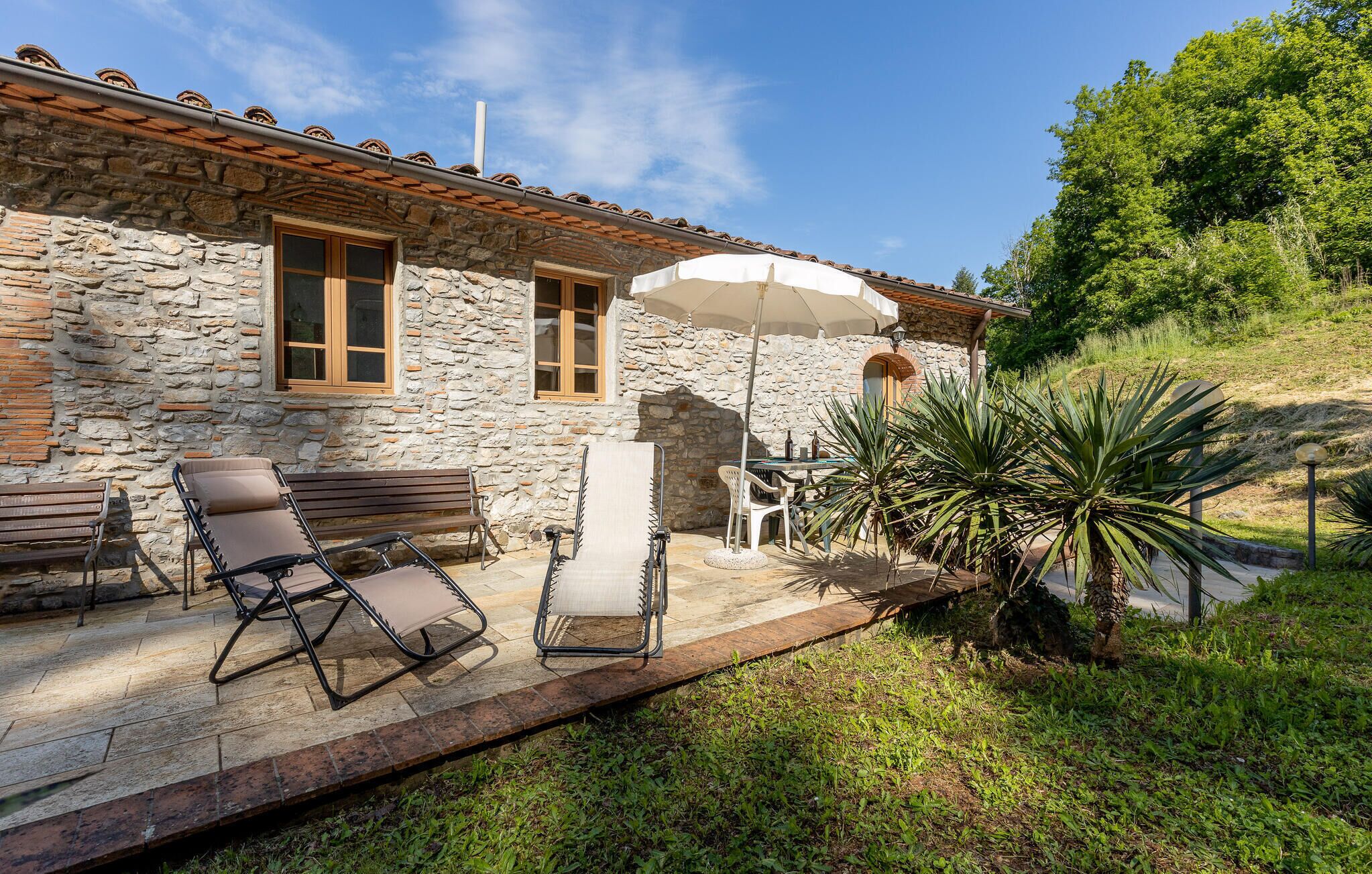 A stone building with a tiled roof, outdoor seating with a table and chairs, and a large umbrella providing shade.