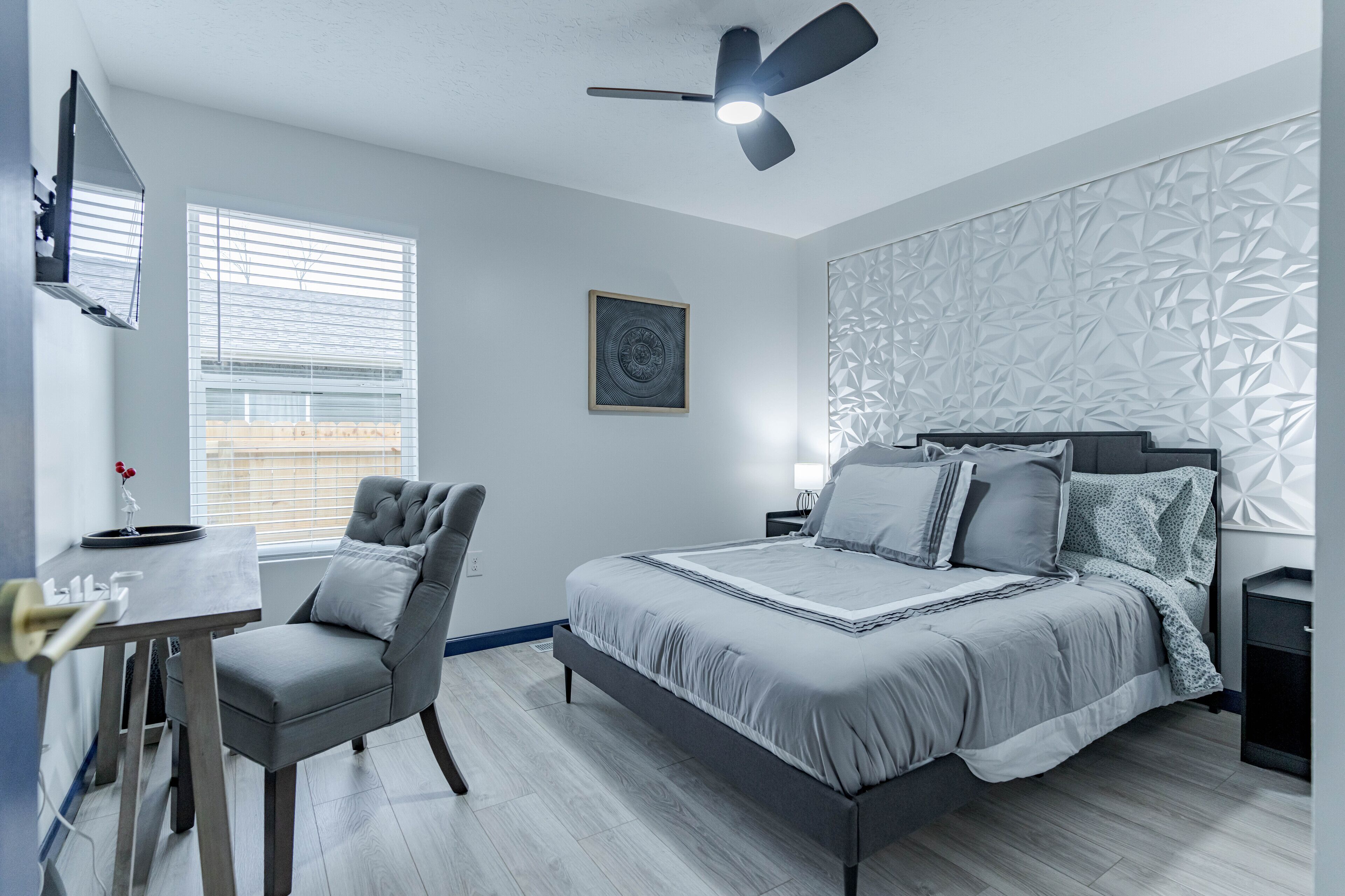 A modern bedroom with a bed, a desk, a chair, a ceiling fan, and a window with blinds.