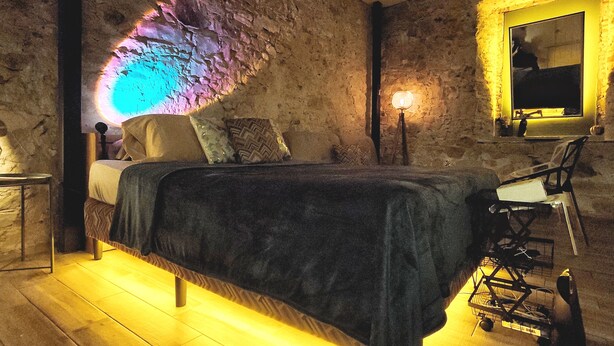 A bedroom with a stone wall, a bed with a dark cover, a bedside table, a lamp, and a chair. - Ocre Studio By Paradox (Panama City)
