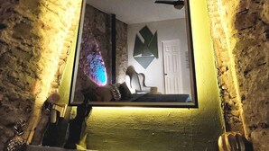 A stone fireplace with a large mirror reflecting a room with a ceiling fan, a chair, and a door. - Ocre Studio By Paradox (Panama City)