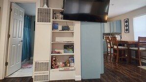 Ein kompakter Wohnraum mit an der Wand montiertem Fernseher, einer Küchenzeile, einem Essbereich mit Holzsesseln und einem Bücherregal mit diversen Gegenständen.