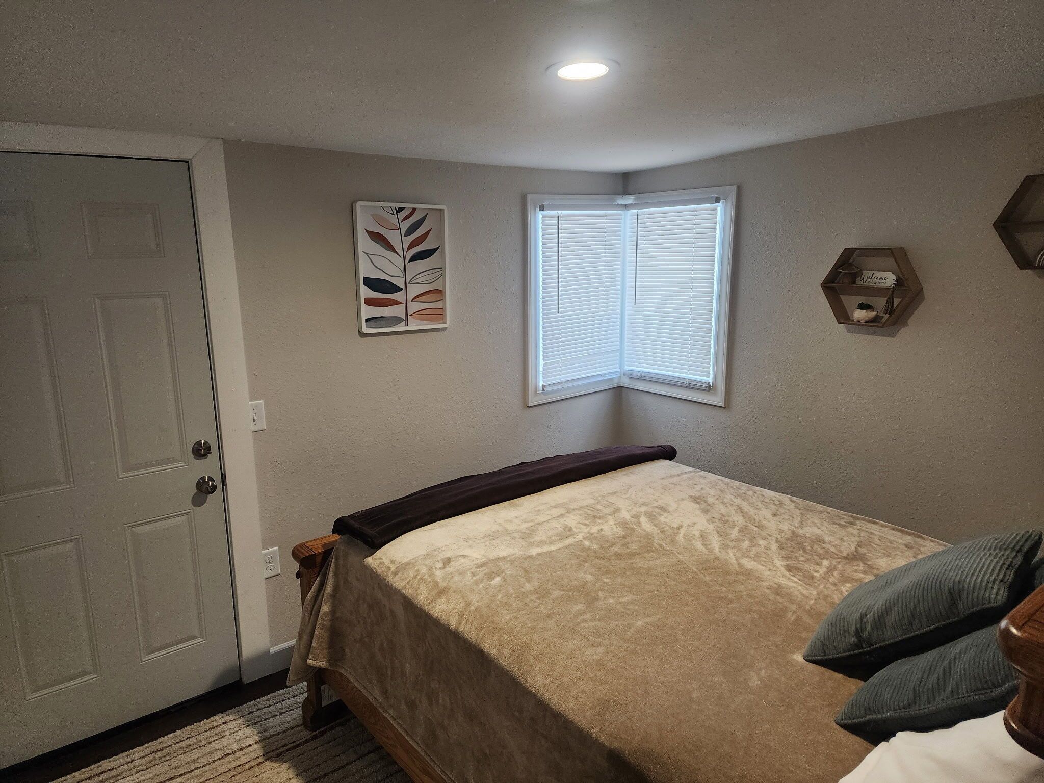 Une chambre à coucher comprenant un lit, une porte, une fenêtre avec des stores, une étagère fixée au mur et un luminaire au plafond.
