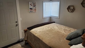 Ein Schlafzimmer mit Bett, einer Tür, einem Fenster mit Jalousien, einem Wandregal und einer Deckenleuchte.