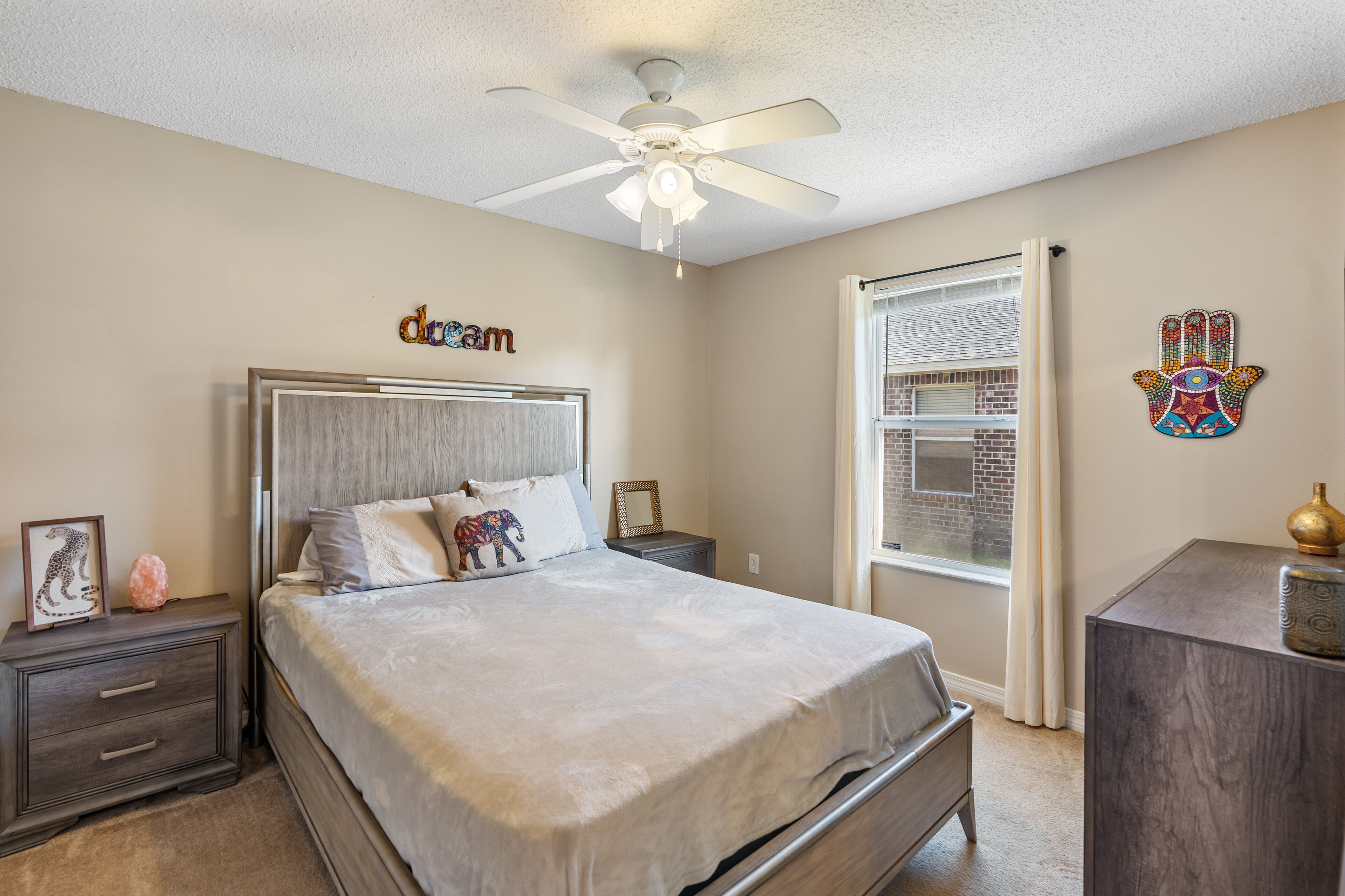 Une chambre à coucher comprenant un lit, des tables de chevet, une commode, un ventilateur de plafond et une fenêtre avec des rideaux.