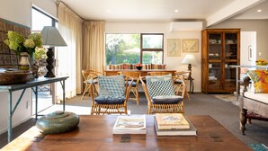 A cozy living room with a wooden coffee table, wicker chairs, and a dining table with chairs. There is a window with curtains, a bookshelf, and a lamp on a side table. - Te Awanga-Zari House- Private&Quiet- 
Children & PETS welcome* (Upon agreement) (Te Awanga)