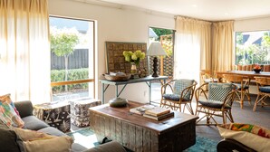 A cozy living room with a sofa, coffee table, and wicker chairs. There is a dining table with chairs and a view of a garden through large windows. - Te Awanga-Zari House- Private&Quiet- 
Children & PETS welcome* (Upon agreement) (Te Awanga)