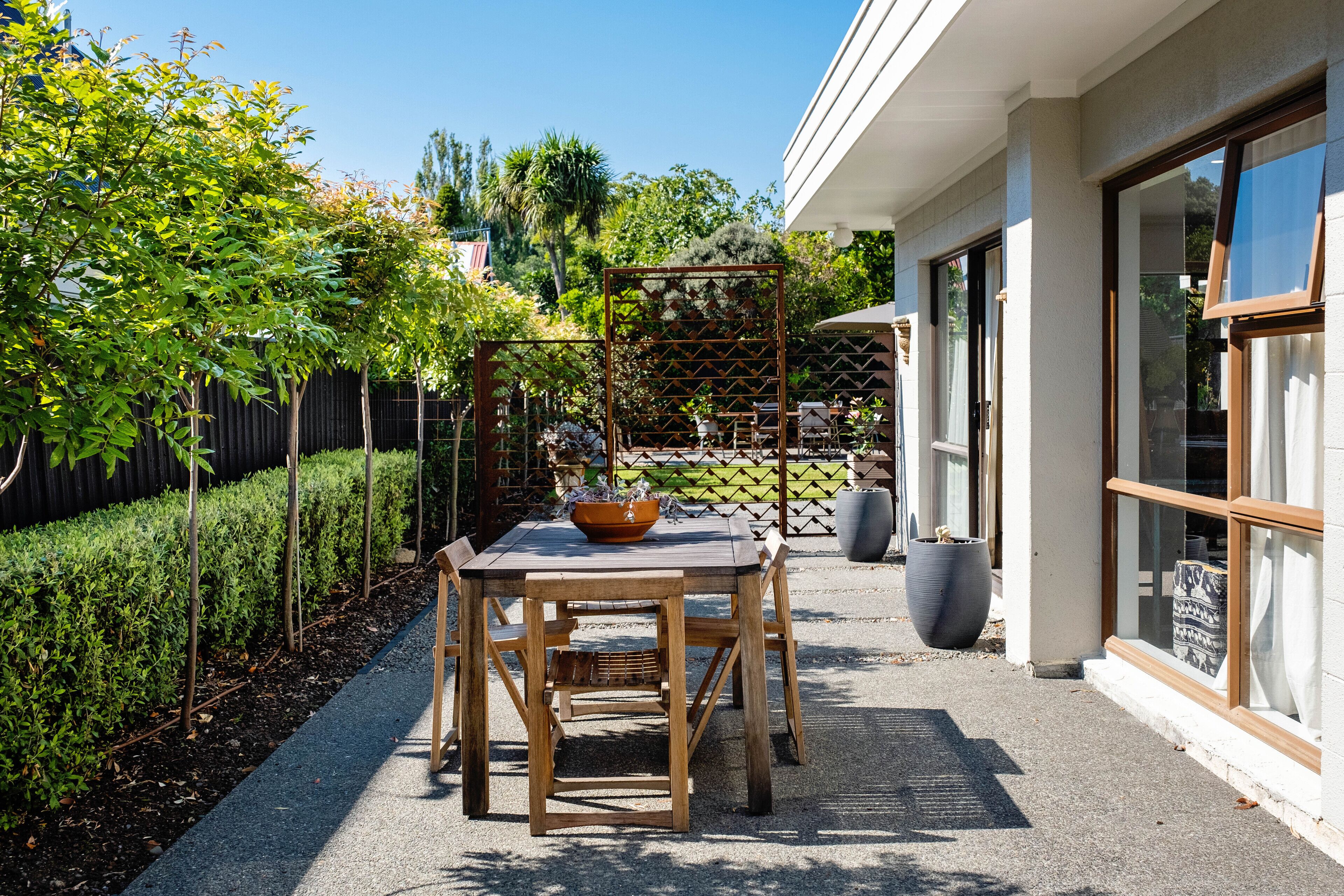 Une terrasse extérieure avec une table et des chaises en bois, entourée de verdure et d’une clôture.