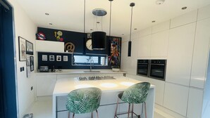 Une cuisine moderne avec des meubles blancs, un îlot central doté de tabourets, un luminaire suspendu noir et une horloge au mur.