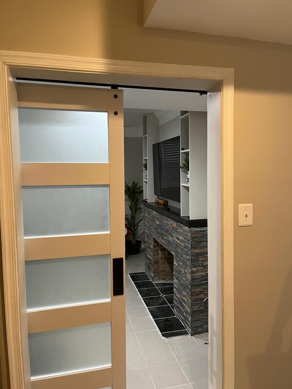 A hallway with a door leading to a living area featuring a fireplace and a television.