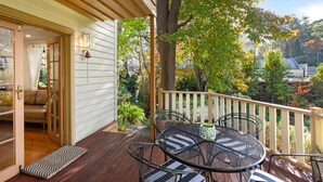 Une terrasse en bois avec une table et des chaises, une porte donnant sur une pièce et une rambarde surplombant un jardin.