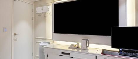 Una habitación de hotel con un televisor de pantalla plana grande montado sobre un escritorio, una cama con cabecera blanca y una pequeña zona de estar con una silla.