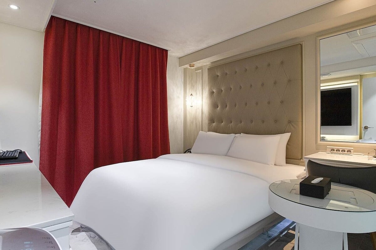 A hotel room with a large bed, a round glass table, a red curtain, a tufted headboard, and a mirror above the table.