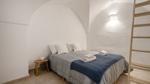A bedroom with a bed, a nightstand, a wall-mounted light, and a wooden ladder. - GuestHost - La Mille - Charming Ostuni Apartment (Ostuni)