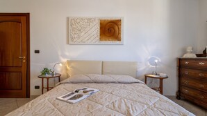 A bedroom with a bed, a nightstand, a dresser, a wall art piece, and a door. - GuestHost - Alghero Sea View Apartment with Balcony! (Alghero)