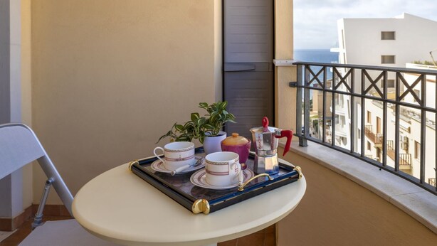 A balcony with a table set for tea, a chair, a potted plant, and a view of buildings and the sea. - GuestHost - Alghero Sea View Apartment with Balcony! (Alghero)