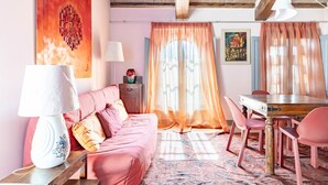 Una habitación con un sofá rosa, una mesa de comedor de madera con sillas, una lámpara y un ventanal con cortinas traslúcidas.