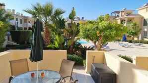 A patio with a round glass table, two chairs, and a trash bin. There are umbrellas and lounge chairs in the background.