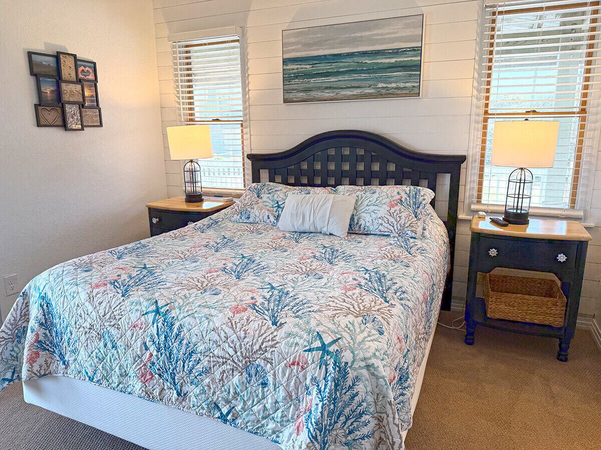 A bedroom with a bed, two bedside tables, a lamp on each table, a wall-mounted picture, and a window with blinds.