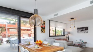 A modern living room with a wooden dining table, white chairs, a grey sofa, and a large window offering a view of the outside.