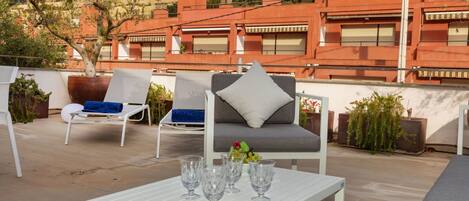 A rooftop terrace with a white table set with wine glasses, a grey sofa with a pillow, and two lounge chairs with blue cushions.