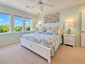 A bedroom with a bed, two bedside tables, a ceiling fan, and a window with a view of greenery.