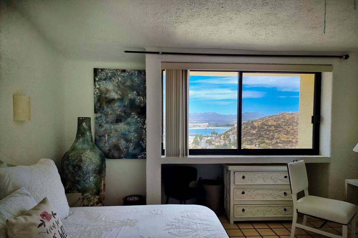 Un dormitorio con cama, una silla, un aparador, una ventana con vistas a un paisaje costero y un cuadro abstracto grande en la pared.