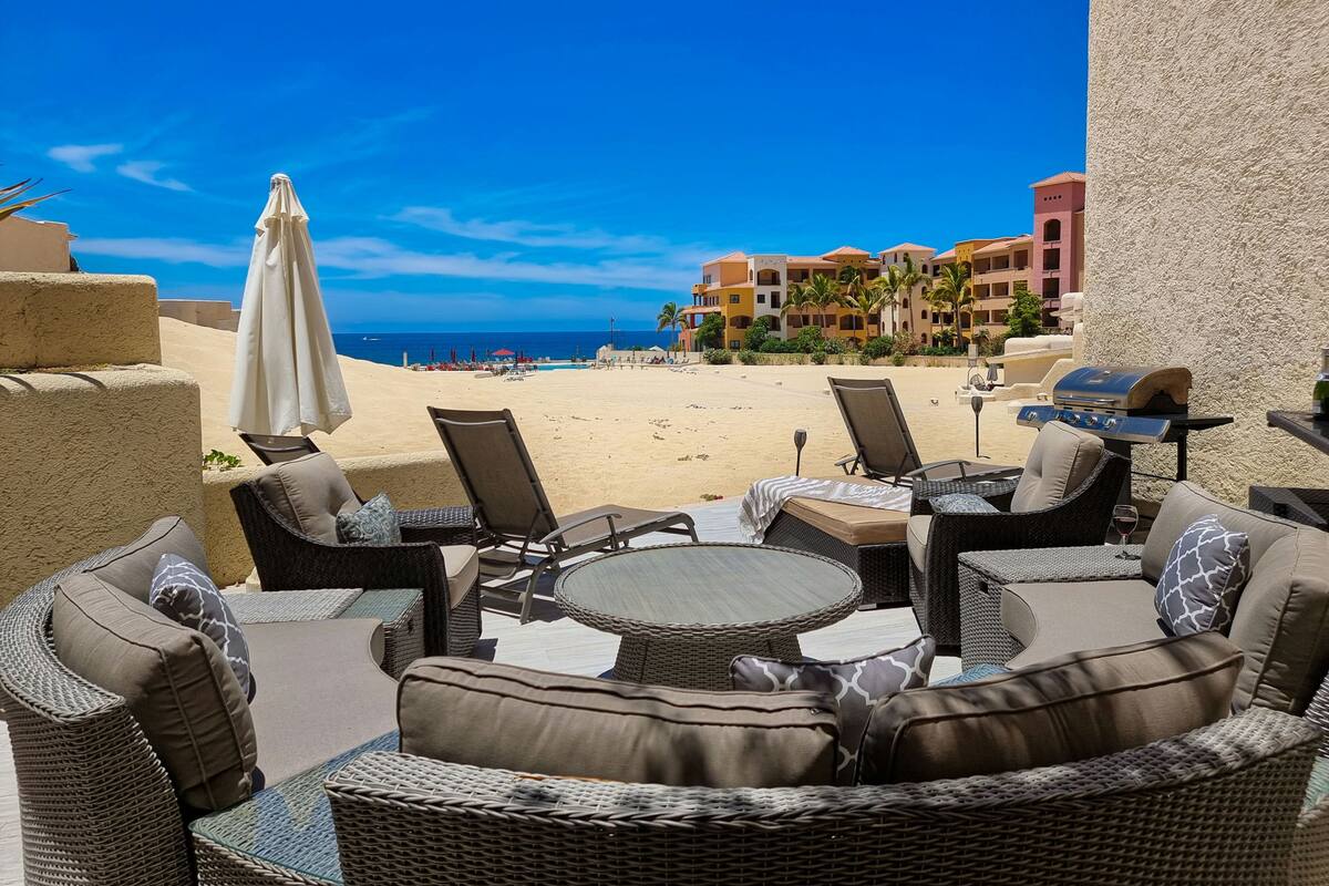 Un patio frente a la playa con mobiliario de mimbre, una parrilla y vistas a la playa y edificios de hotel.