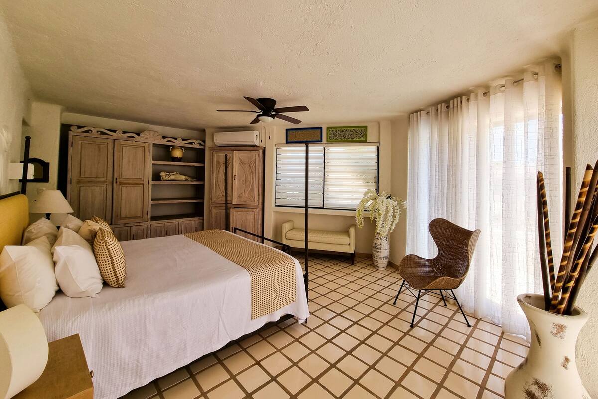 Un dormitorio con cama, una silla, ventilador de techo y ventana con cortinas.