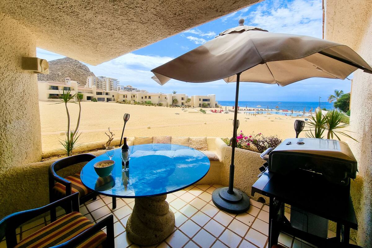 Un patio con una mesa redonda, sillas, una parrilla y vista a la playa.