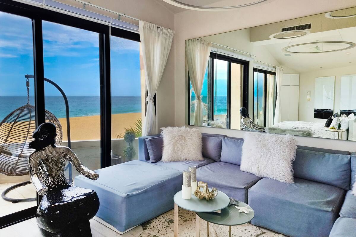 Habitación de hotel moderna con un amplio ventanal que ofrece vista al mar, una silla colgante, un sofá y una mesita con adornos.