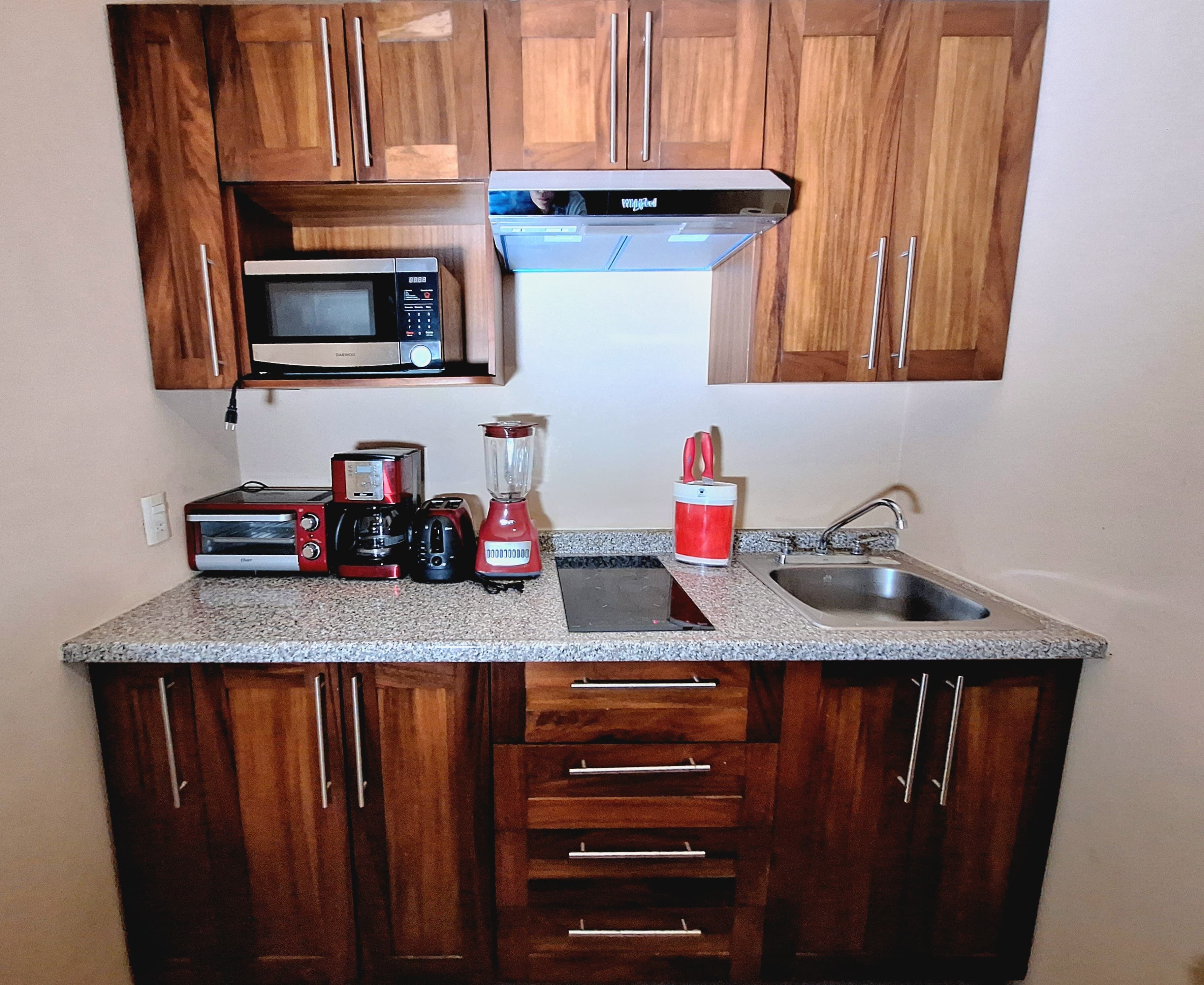 A compact kitchen with wooden cabinets, a microwave, a toaster, a coffee maker, a blender, and a sink.